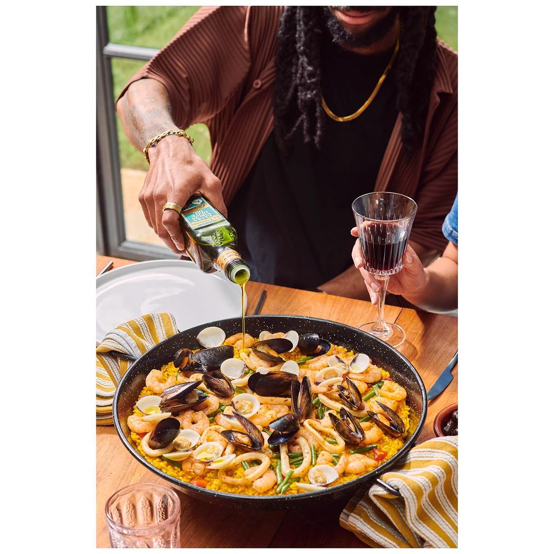 Overhead shot of paella being drizzled with olive oil by a man at an outdoor table setting alongside a wine glass.
