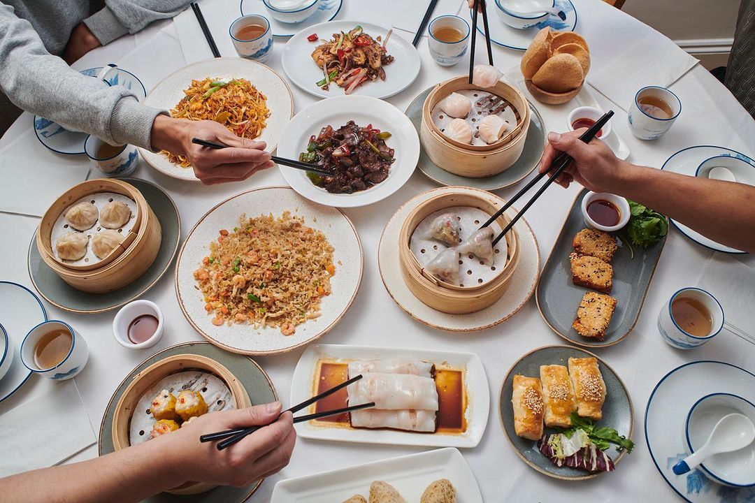 Overhead shot of a group enjoying a diverse spread of Asian cuisine on a round table, featuring various dumplings, noodles, and fried dishes.