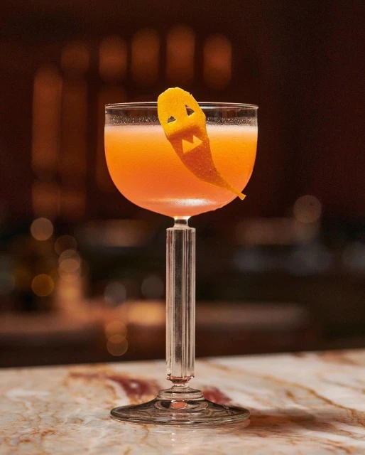 Halloween cocktail in coupe glass with a carved orange peel garnish on a marble countertop, against a blurred background.