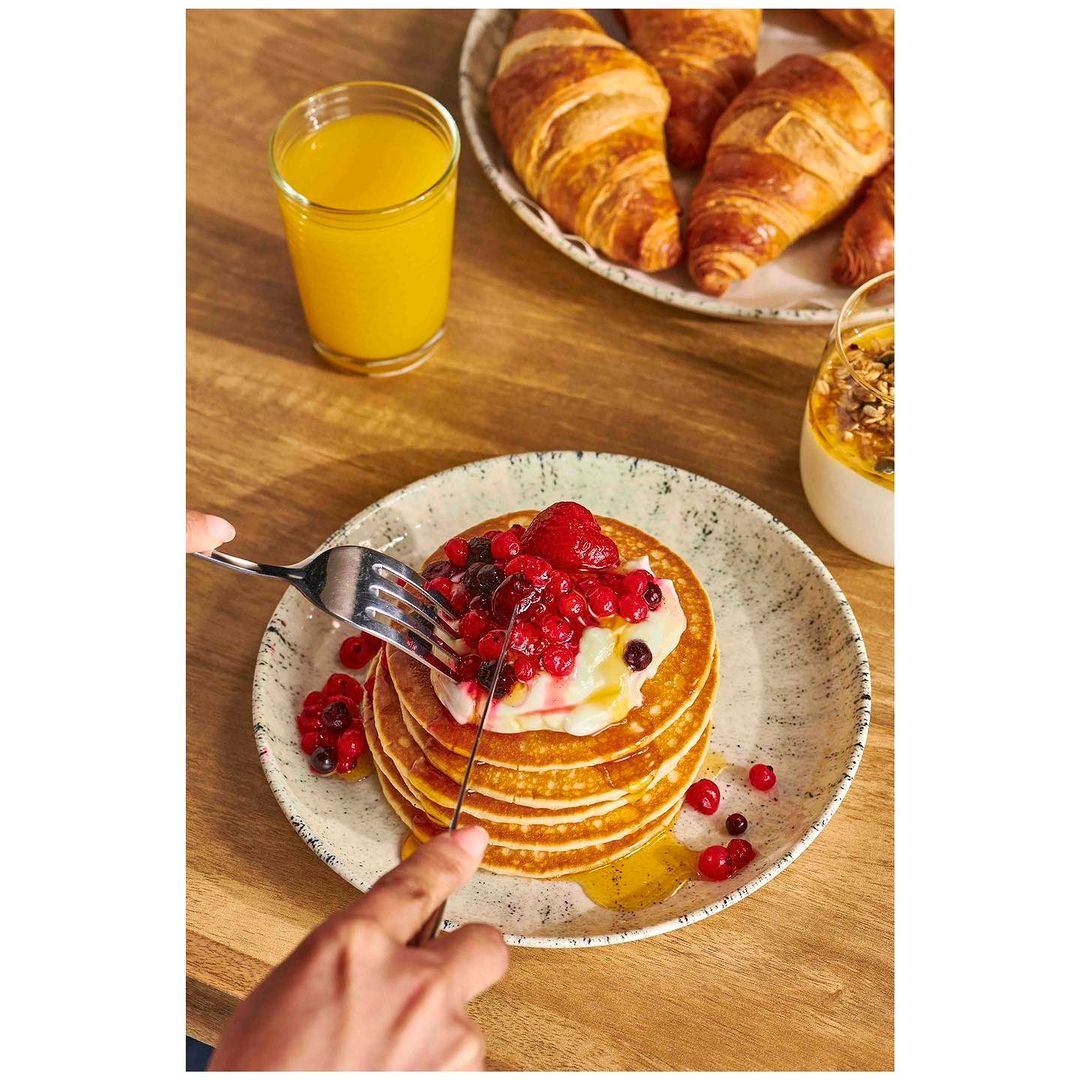 Overhead view of pancakes with berries, yogurt, and syrup being cut with cutlery, alongside juice and croissants on a wooden table.