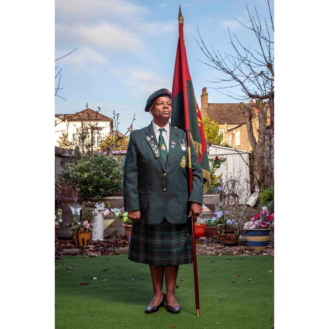 Mature African American man in green uniform and kilt stands holding a flag in a garden setting with a house in the background.