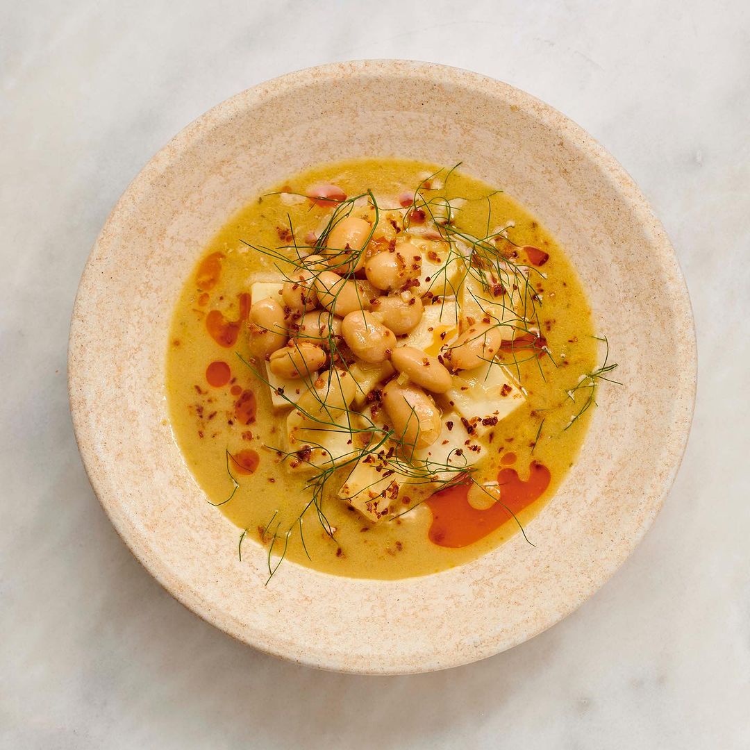 Overhead shot of a tan ceramic bowl filled with white beans, chunks of white cheese, and a yellow sauce, garnished with dill.