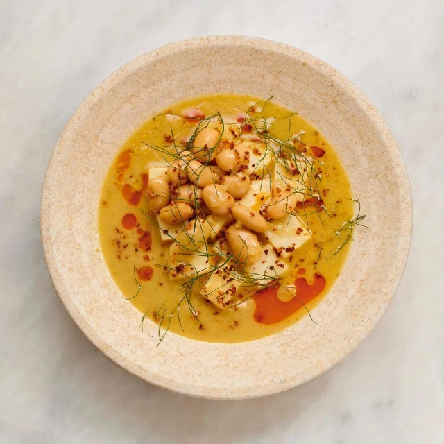 Overhead shot of a tan ceramic bowl filled with white beans, chunks of white cheese, and a yellow sauce, garnished with dill.