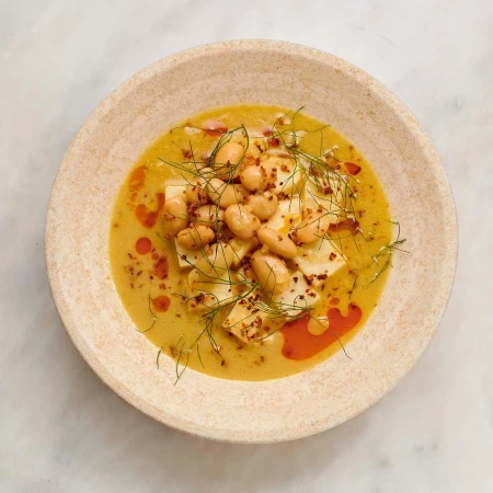 Overhead shot of a tan ceramic bowl filled with white beans, chunks of white cheese, and a yellow sauce, garnished with dill.
