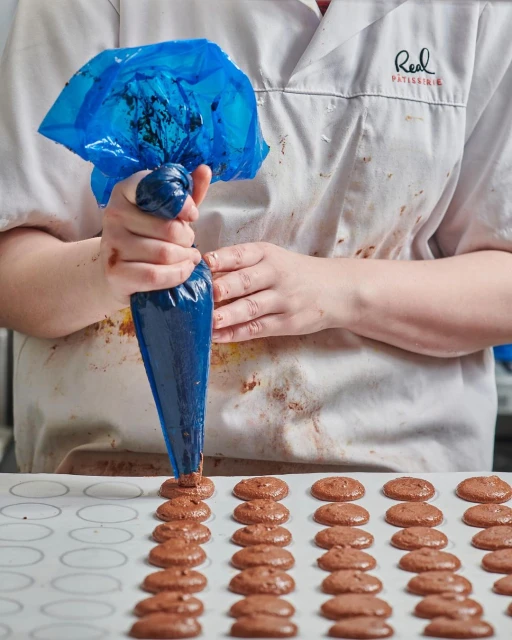 Pastry chef using a piping bag to fill rows of chocolate macarons on a white tray in a commercial kitchen. Close-up shot.
