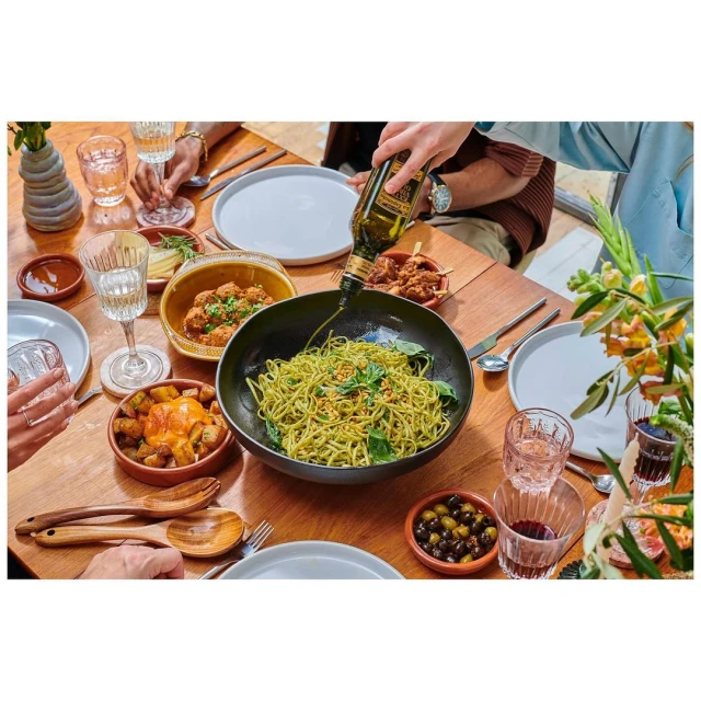 Overhead shot of green pasta dish on a wood table with various side dishes, olive oil being poured, and several people eating.