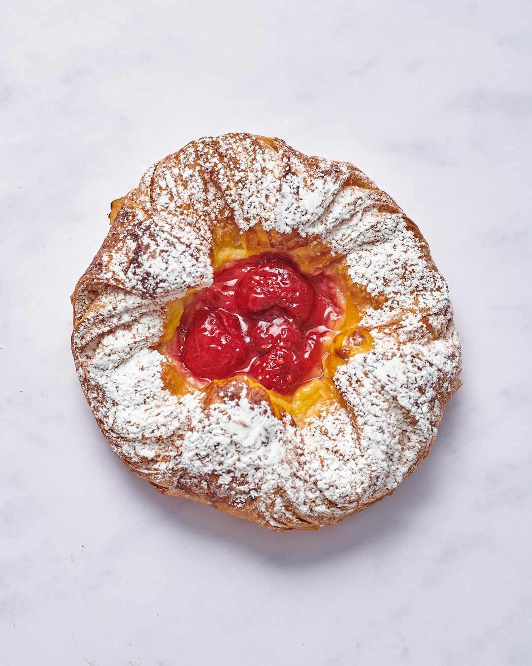 Overhead shot of a freshly baked pastry filled with a red fruit filling and dusted with powdered sugar on a light marble surface.