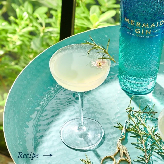 A light yellow cocktail in a coupe glass sits on a teal hammered metal table next to a bottle of gin, sprigs of rosemary, and shears.