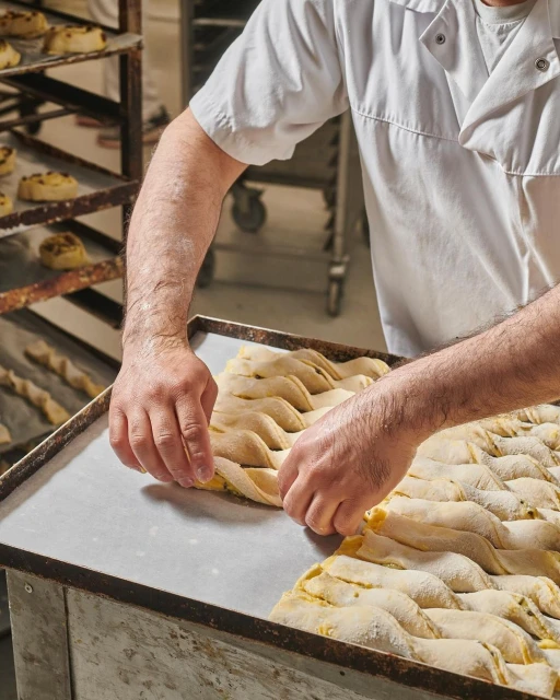 Baker shaping raw dough on a metal sheet in a commercial kitchen, with rows of baking racks in the background, under bright lighting.