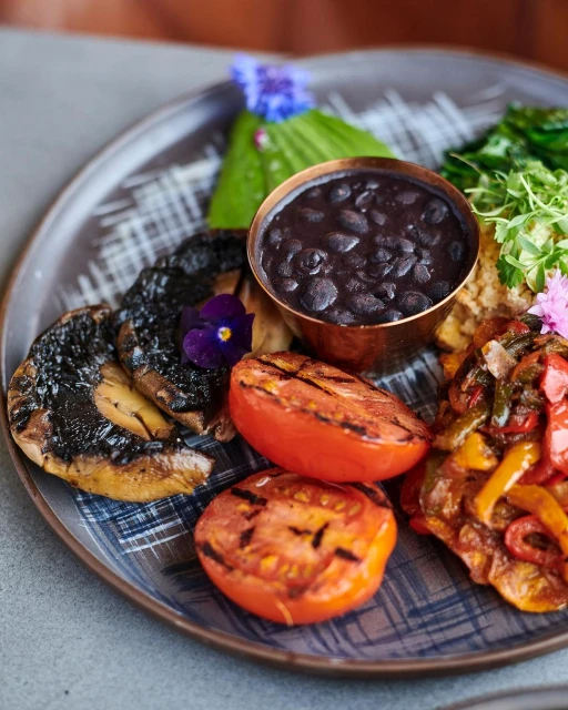 A flavorful plate featuring grilled tomatoes, mushrooms, black beans in a copper bowl, colorful sauteed bell peppers, and fresh herbs.