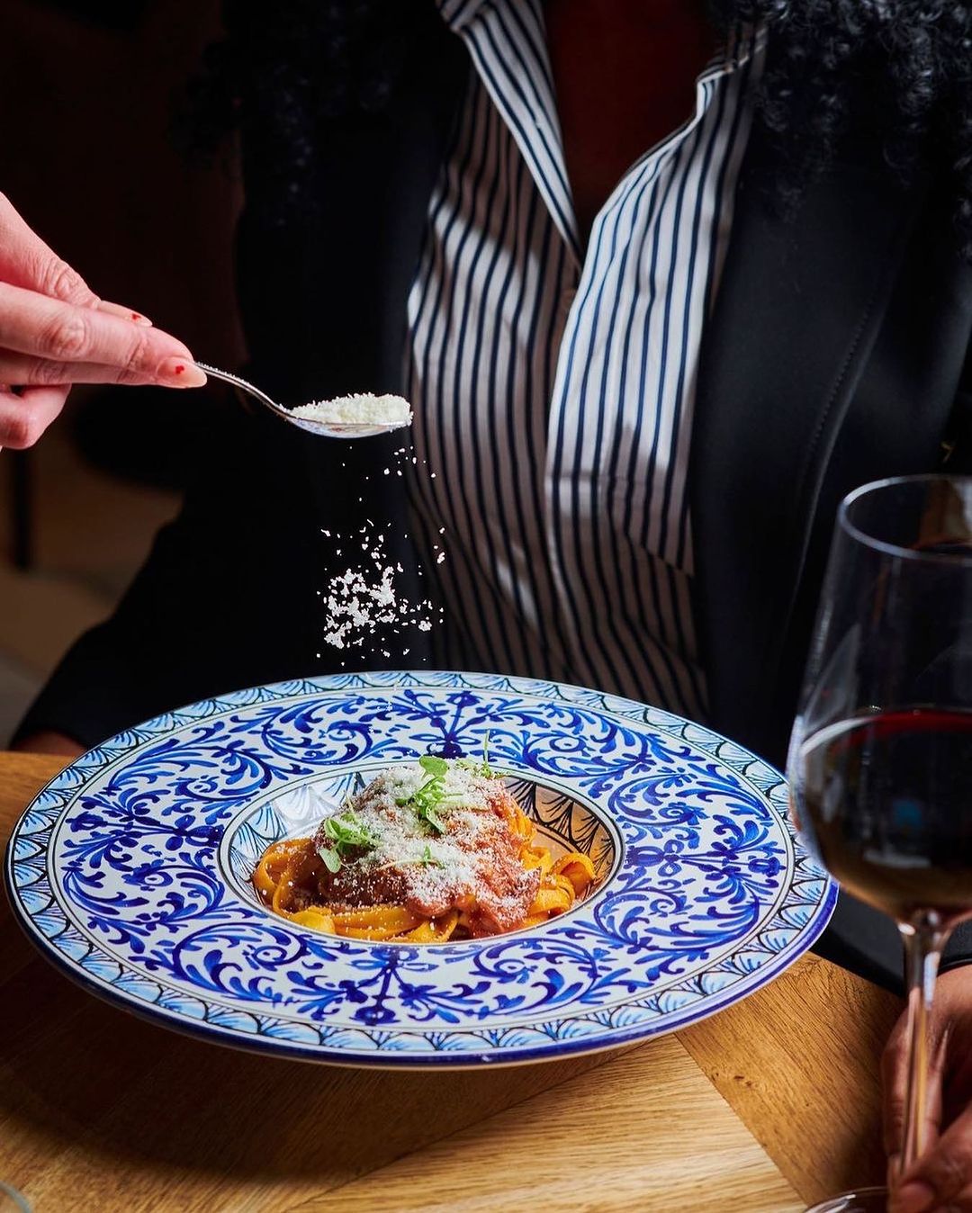 Overhead shot of ornate plate of pasta with a red sauce, herbs, and grated cheese, a spoonful of cheese and wine glass visible.