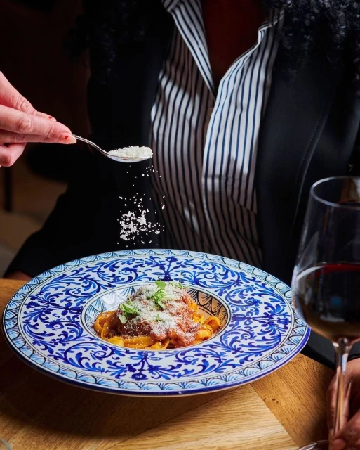 Overhead shot of ornate plate of pasta with a red sauce, herbs, and grated cheese, a spoonful of cheese and wine glass visible.
