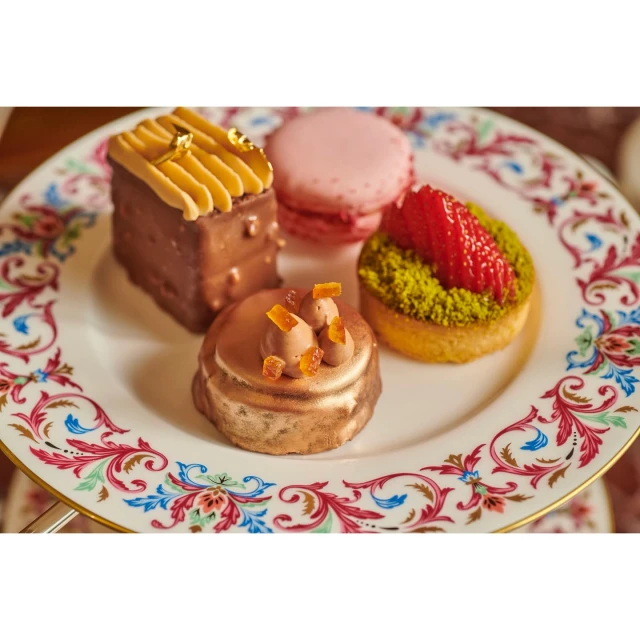 Close-up of an ornate plate with four small cakes, including a chocolate cube with caramel, a pink macaron, and a strawberry tart.