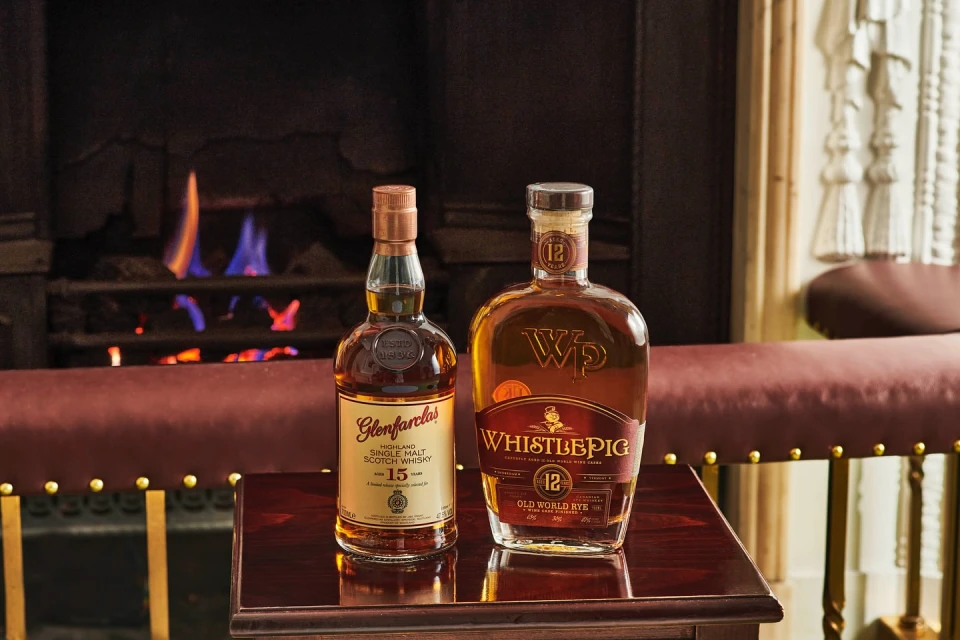 Two bottles of aged whiskey stand on a table in front of a fireplace, the warm tones of the liquor complementing the cozy setting.
