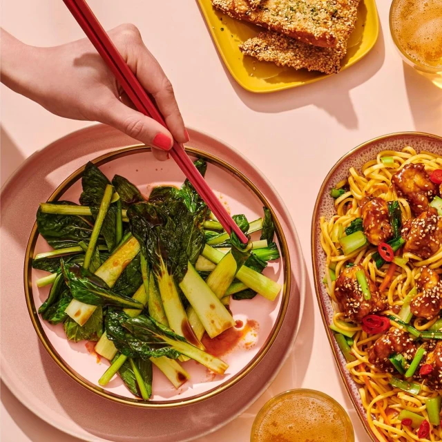 Overhead shot of a plate of bok choy being eaten with chopsticks next to noodles and appetizers; tabletop food and drink presentation.