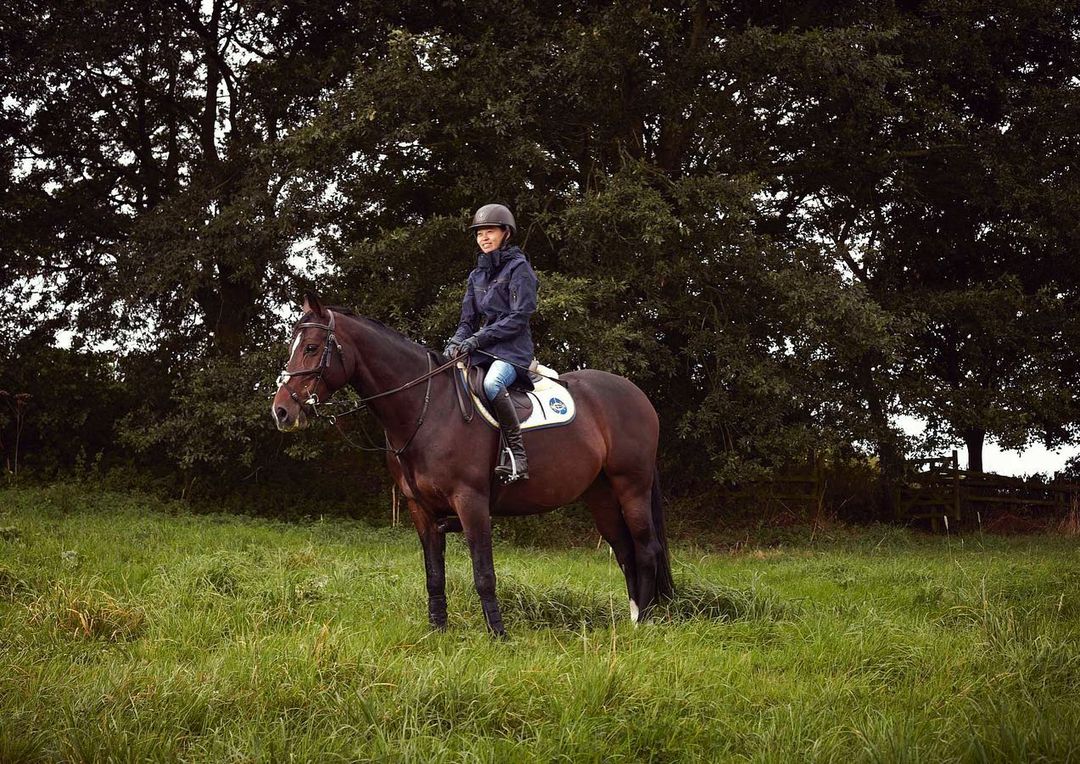 Woman riding a brown horse through a grassy field, wearing a helmet and riding gear, with a tree-filled background.