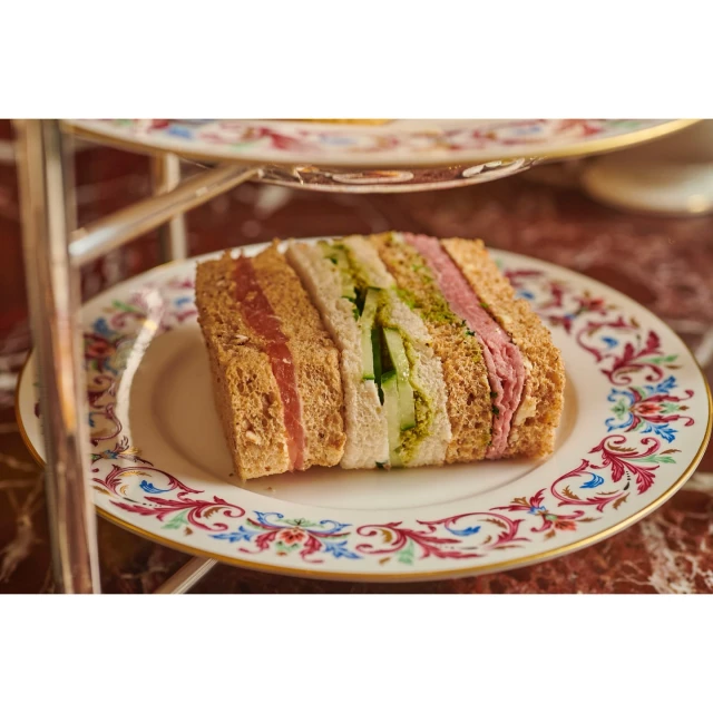 Afternoon tea: stacked sandwiches on a floral plate atop a multi-tiered stand. Focus on fresh, colourful sandwiches.
