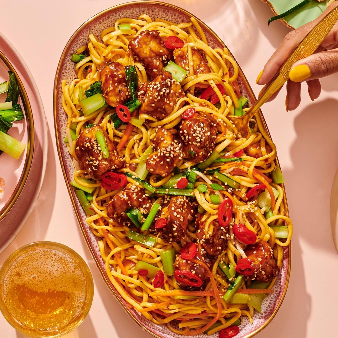 Overhead shot of noodles with glazed chicken on an oval platter, garnished with vegetables, and a golden fork lifting the food.