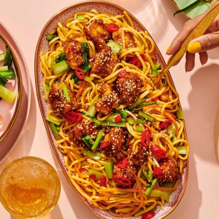 Overhead shot of noodles with glazed chicken on an oval platter, garnished with vegetables, and a golden fork lifting the food.