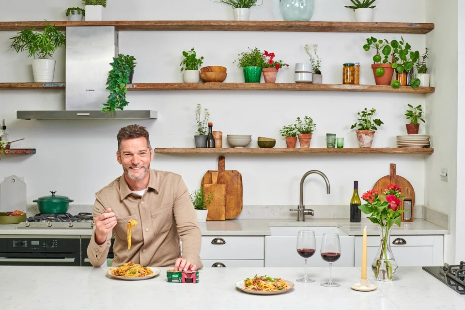 Adult male smiles in a kitchen with pasta, wine, flowers, and plants on shelves and the counter creating a bright, inviting atmosphere.