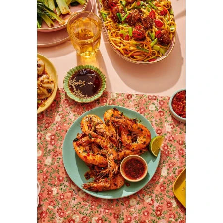 Overhead shot of shrimp on a plate with sauces, noodles with meatballs, and a glass of white wine on a pink floral tablecloth.