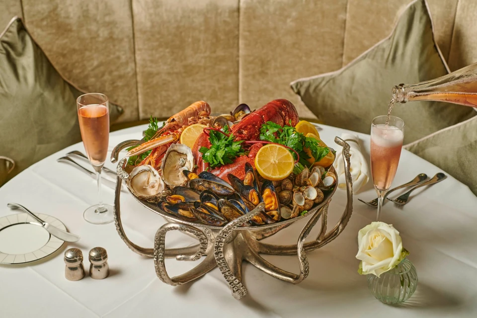 Seafood platter with lobster, oysters, and mussels is displayed on a silver octopus stand with champagne on a white tablecloth.