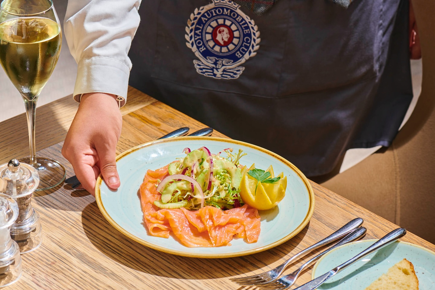 Waiter holding a plate of salmon salad, complete with lemon and greens, next to a glass of white wine on a restaurant table.