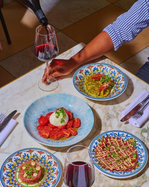 Overhead shot of four colorful plates of Italian food on a marble table, including pasta, tomatoes with burrata, and steak tartare, with wine.
