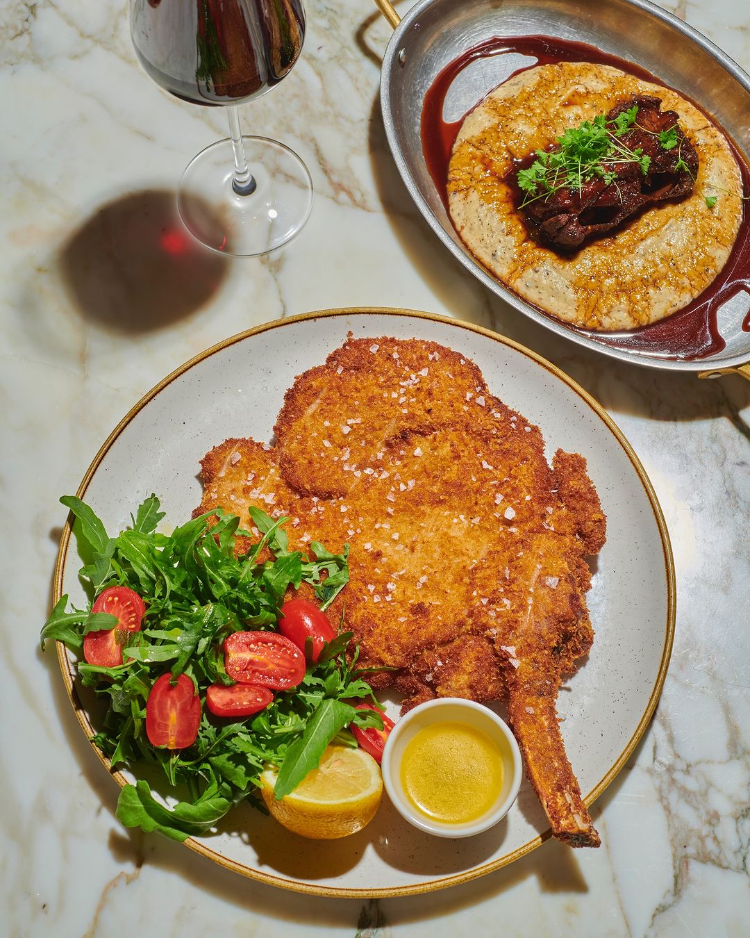 Overhead shot of golden schnitzel with salad, lemon, and mustard on a white marble table, with wine glass and other dishes.