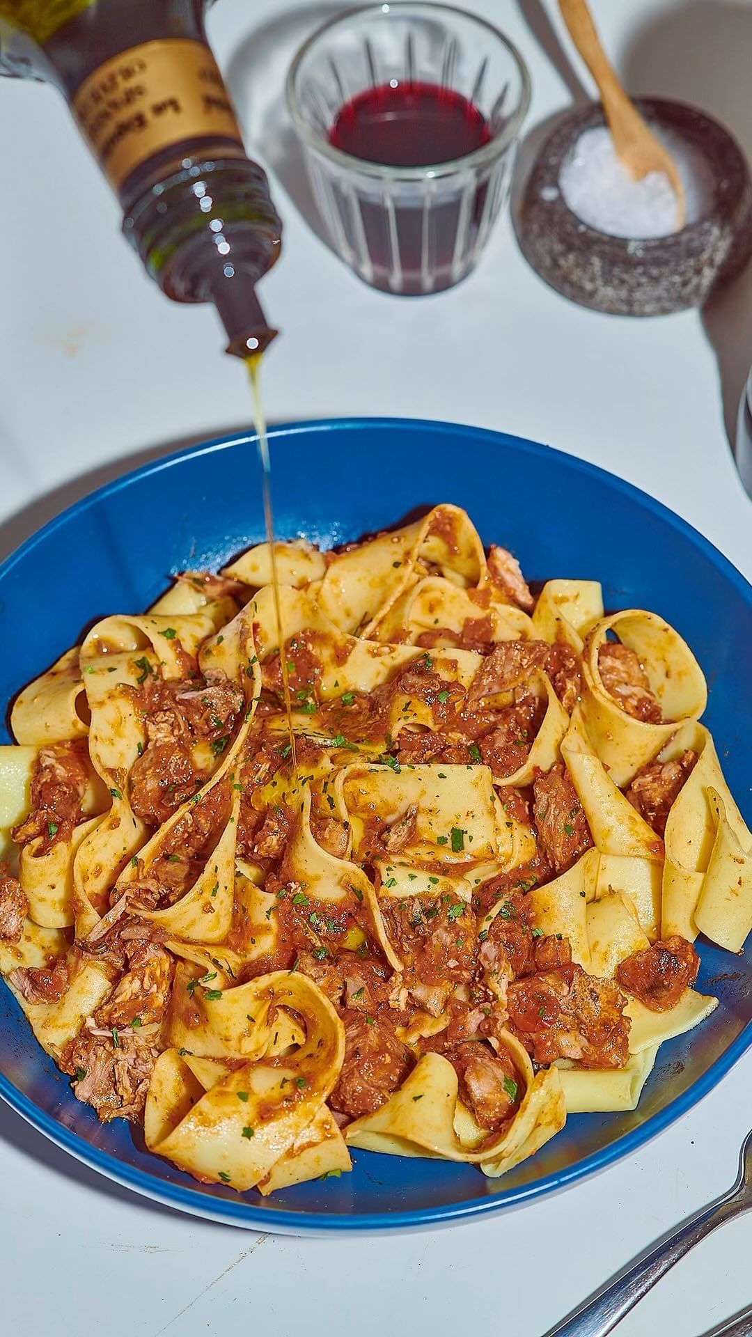 Overhead shot of large pasta dish with meat sauce, drizzled with oil, alongside a bottle, salt cellar, and glass of wine.
