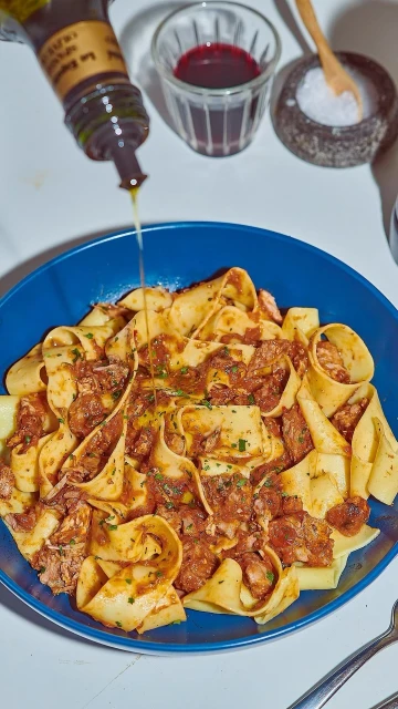 Overhead shot of large pasta dish with meat sauce, drizzled with oil, alongside a bottle, salt cellar, and glass of wine.