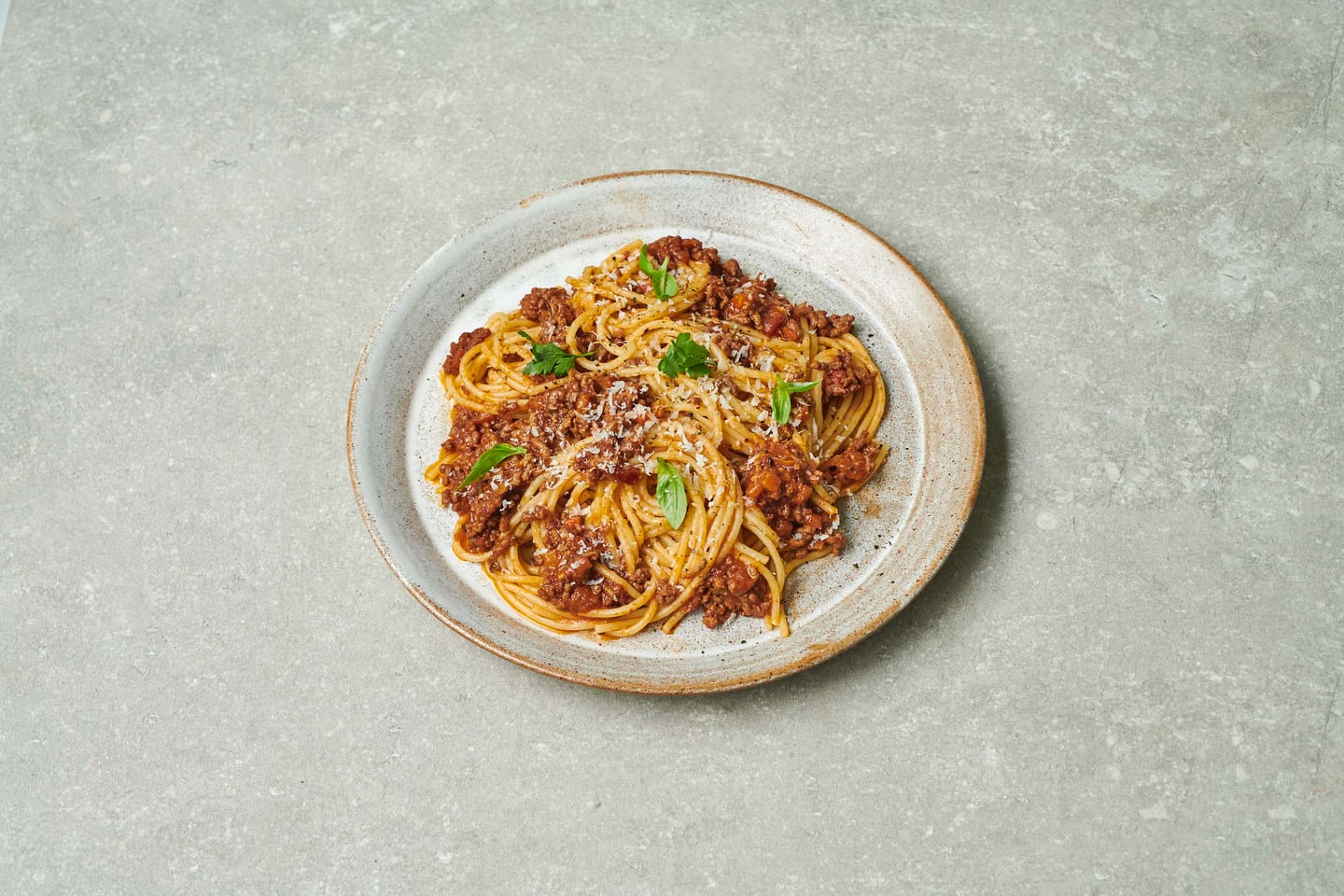 Overhead shot of spaghetti bolognese on a gray plate, topped with shredded cheese and basil, placed on a textured gray surface.