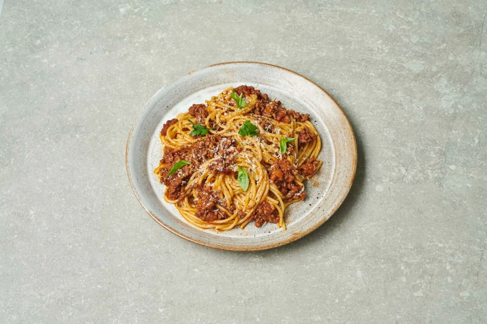Overhead shot of spaghetti bolognese on a gray plate, topped with shredded cheese and basil, placed on a textured gray surface.