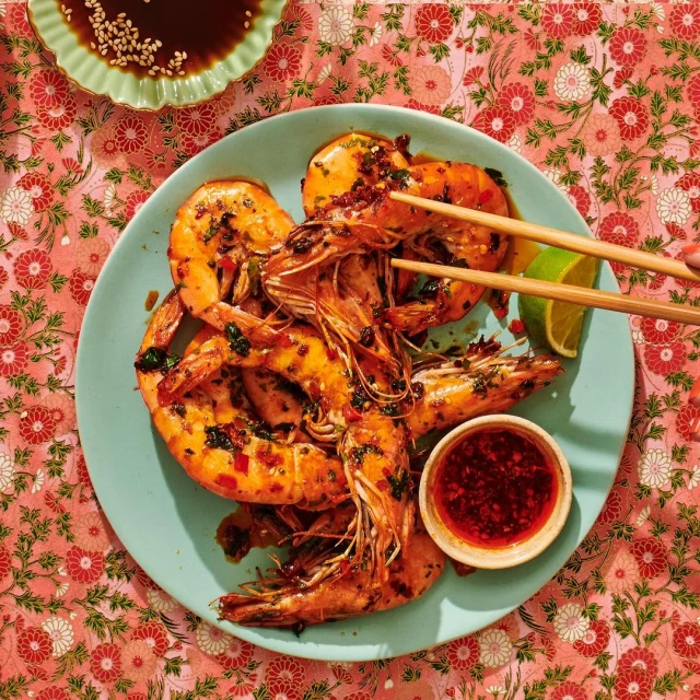 Overhead food shot: cooked shrimps seasoned with spices on a blue plate, chopsticks, chili sauce, lime and a floral tablecloth.