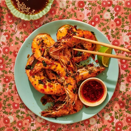 Overhead food shot: cooked shrimps seasoned with spices on a blue plate, chopsticks, chili sauce, lime and a floral tablecloth.
