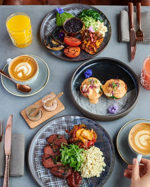Overhead shot of three plates with breakfast food, orange juice, and coffee, arranged on a gray table, featuring vibrant colors and textures.