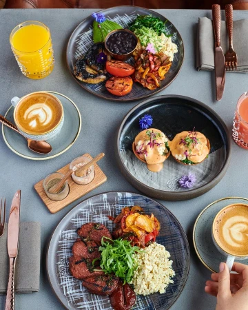 Overhead shot of three plates with breakfast food, orange juice, and coffee, arranged on a gray table, featuring vibrant colors and textures.