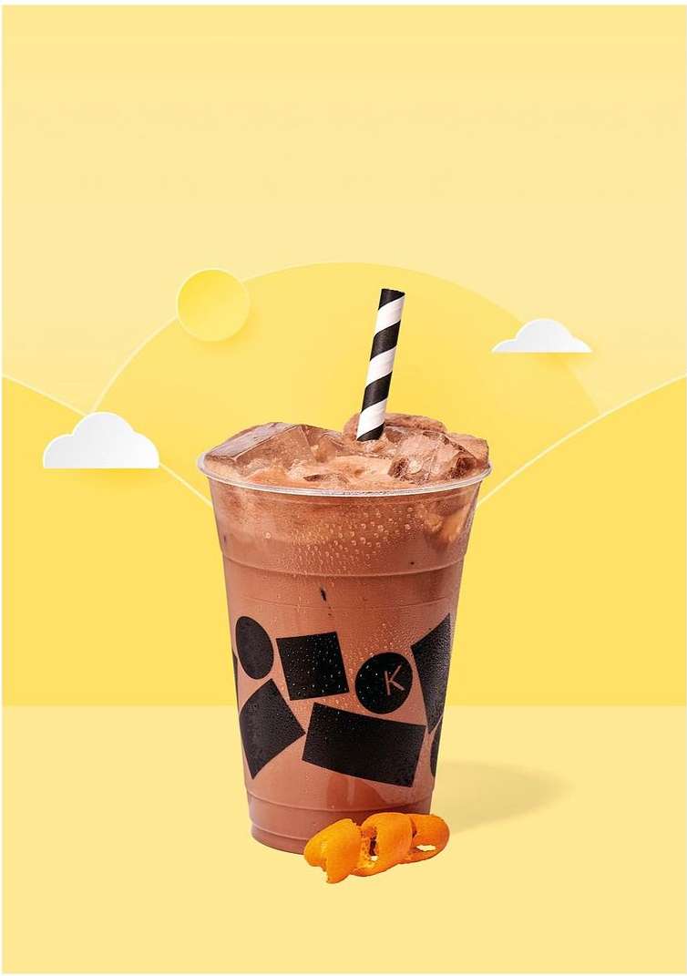 Refreshing iced chocolate drink in a clear plastic cup with a striped straw, set against a sunny yellow backdrop with paper-cutout decorations.