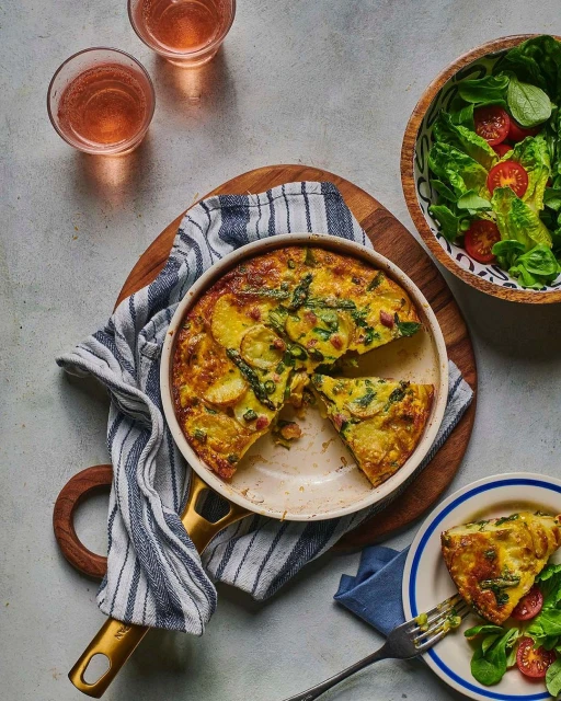 Overhead shot of a frittata in a skillet, plated slice, salad, and drinks creating an inviting food and beverage display.