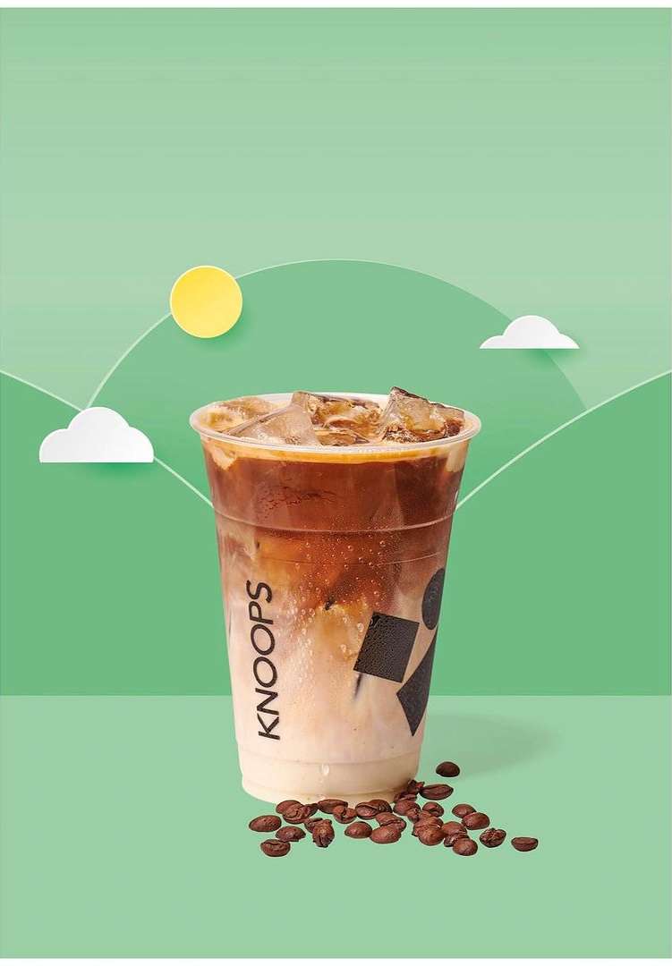 Iced latte in a clear plastic cup with coffee beans scattered on a light green surface against a backdrop of abstract hills.