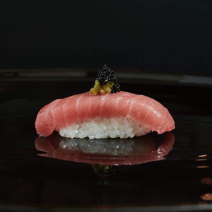 Close-up of nigiri sushi with tuna, rice, green wasabi, and black caviar on a shiny black plate reflecting the composition.