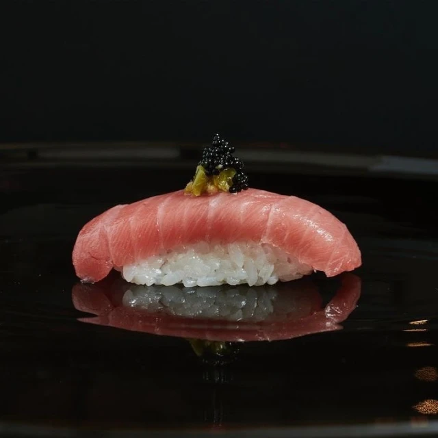 Close-up of nigiri sushi with tuna, rice, green wasabi, and black caviar on a shiny black plate reflecting the composition.