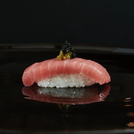 Close-up of nigiri sushi with tuna, rice, green wasabi, and black caviar on a shiny black plate reflecting the composition.