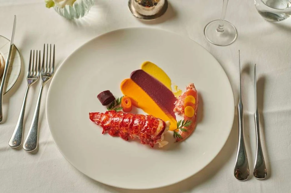 Gourmet lobster dish on a white plate with colorful sauces and minimalist silverware, presented on a white tablecloth.