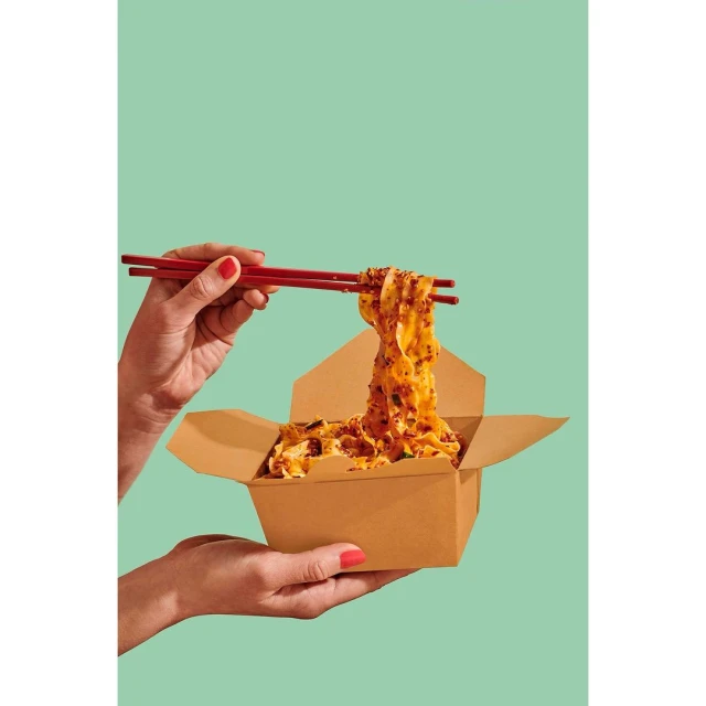 Overhead shot: Hands using red chopsticks lifting noodles from a cardboard takeout container, set against a solid aqua-green background.
