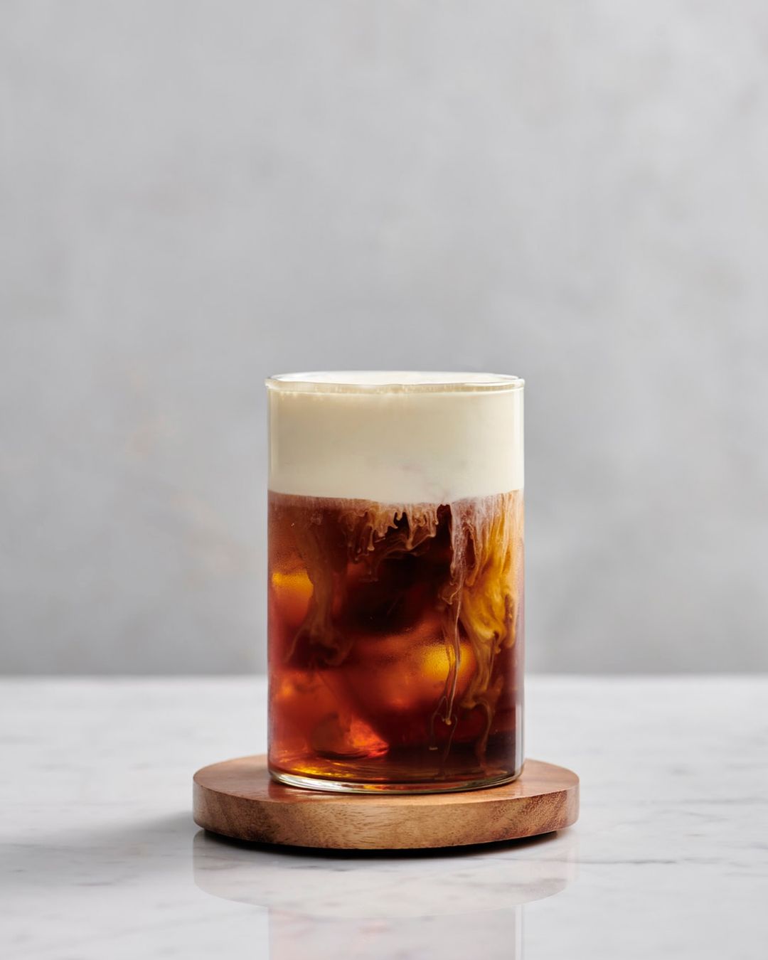 A glass of iced coffee topped with a layer of cream, sitting on a wooden coaster, creating a visually appealing beverage.