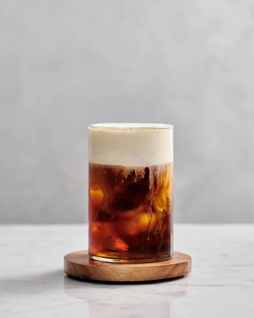 A glass of iced coffee topped with a layer of cream, sitting on a wooden coaster, creating a visually appealing beverage.