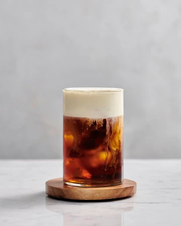 A glass of iced coffee topped with a layer of cream, sitting on a wooden coaster, creating a visually appealing beverage.