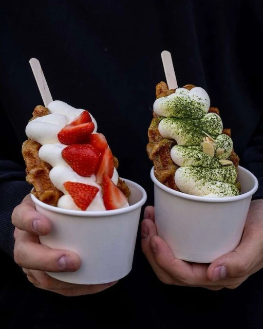 Two hands hold paper cups filled with waffle ice cream, topped with fresh strawberries and matcha powder against a dark background.