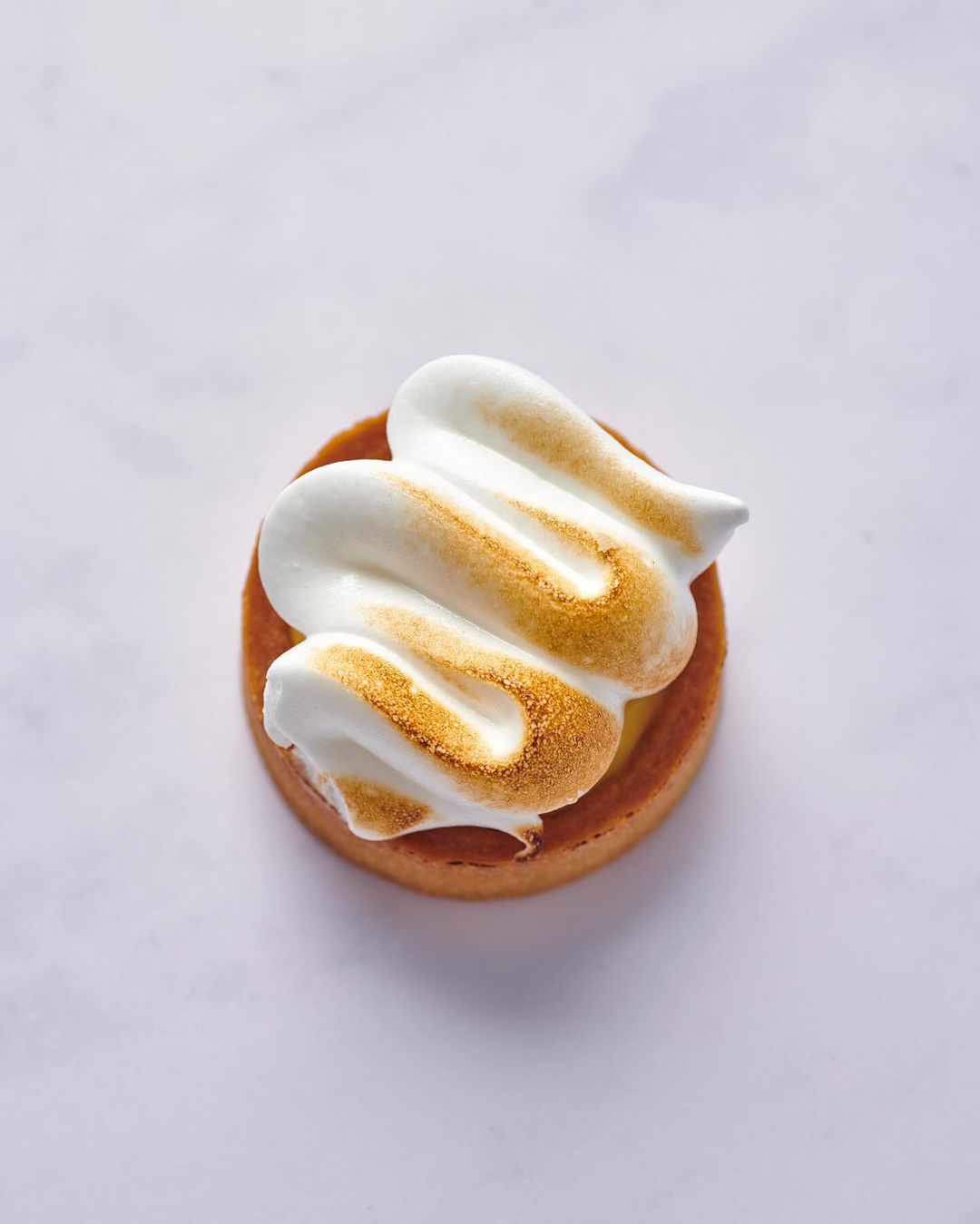 Overhead shot of a single lemon meringue tart on a white surface, showcasing the pastry crust and the torched meringue topping.